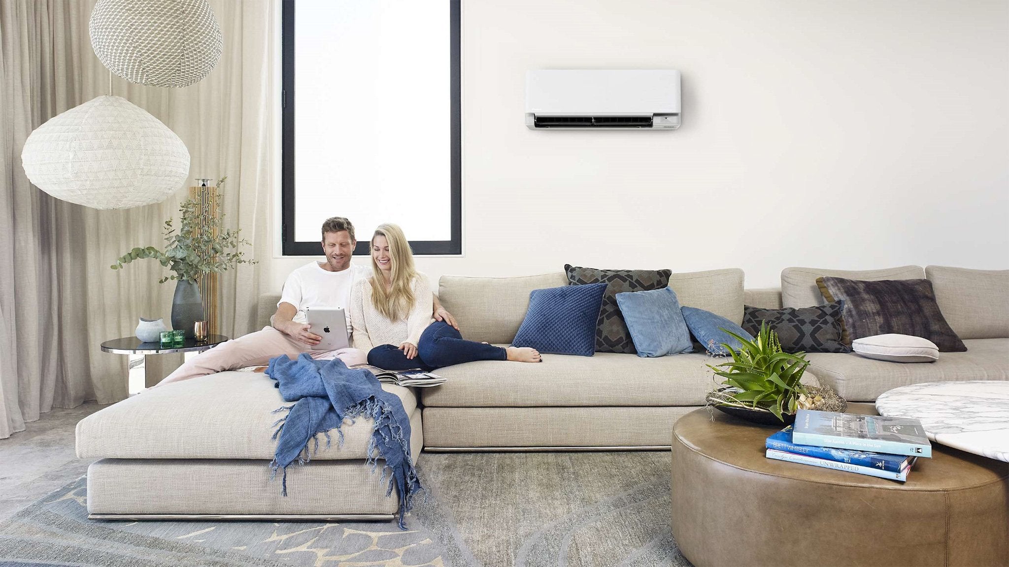 Couple sitting on a beige sectional sofa in a modern living room with a wall-mounted air conditioner.