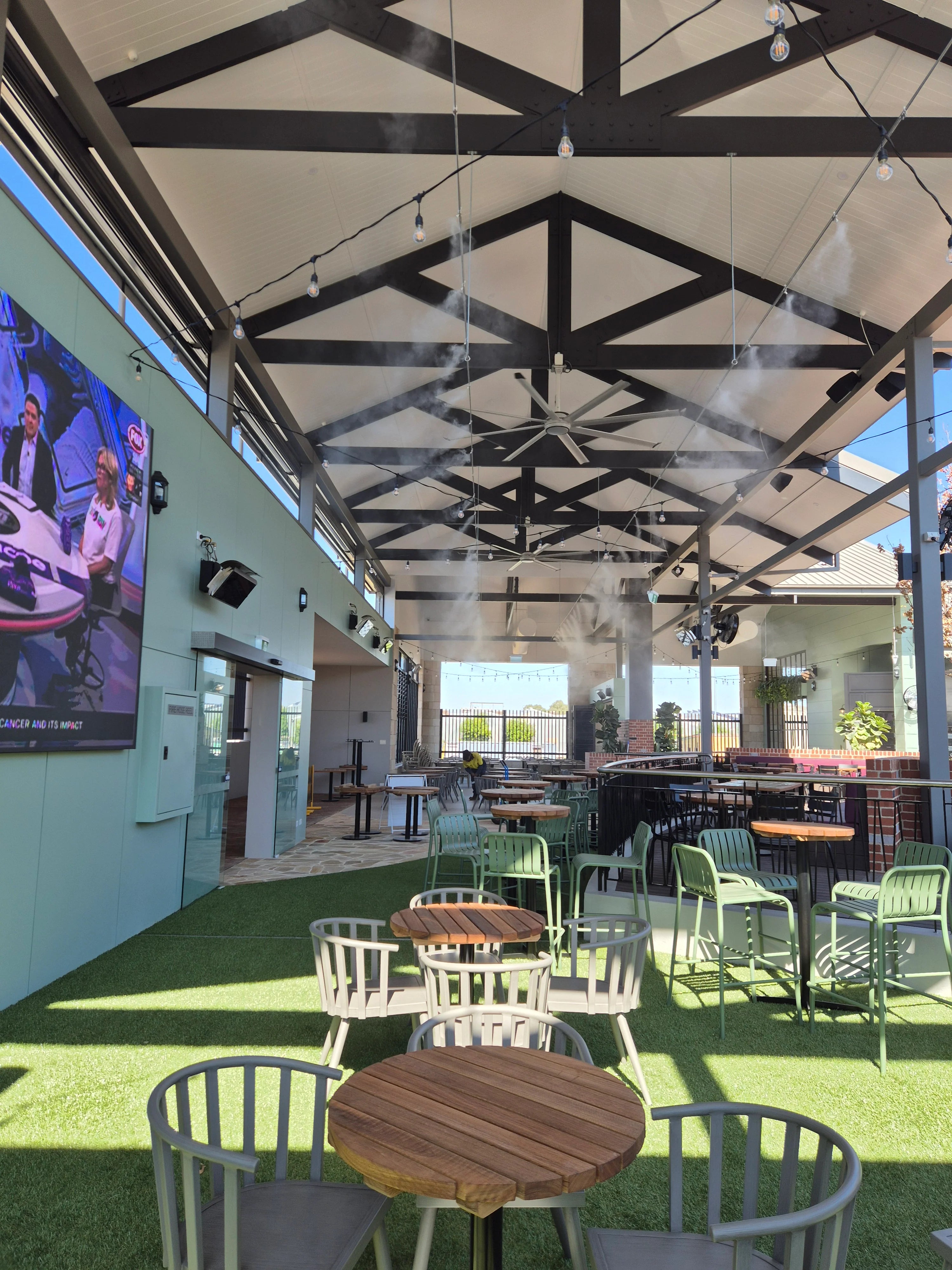 Outdoor seating area with tables and chairs under a large open roof structure with spray cooling