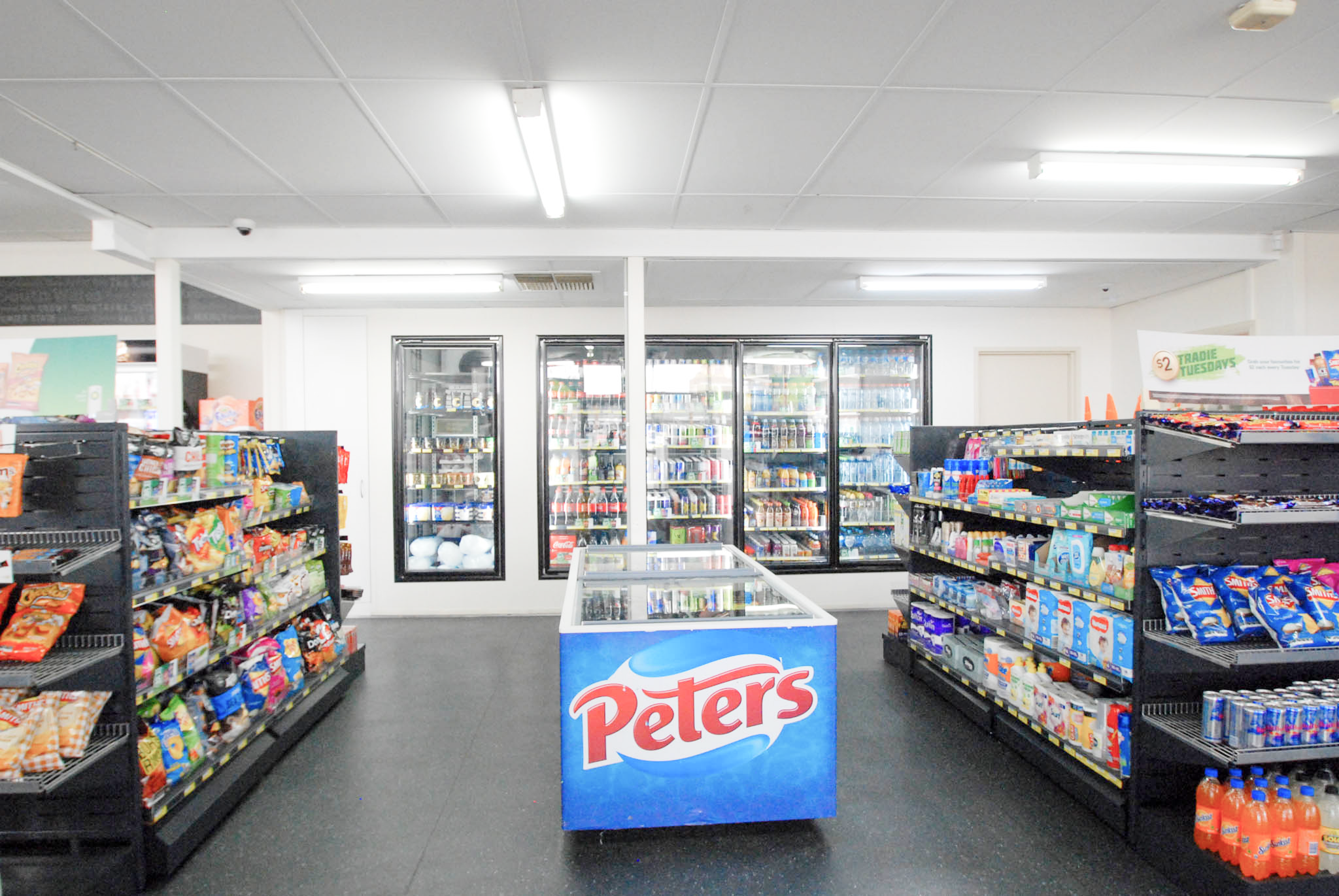 Supermarket interior with shelves stocked with products and a 'Peters' branded cooler.